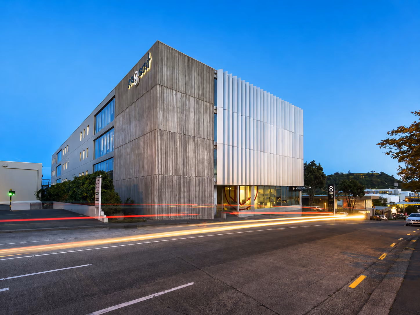 Modern multi-story office building with concrete and white panel facade at dusk with light trails on the street.