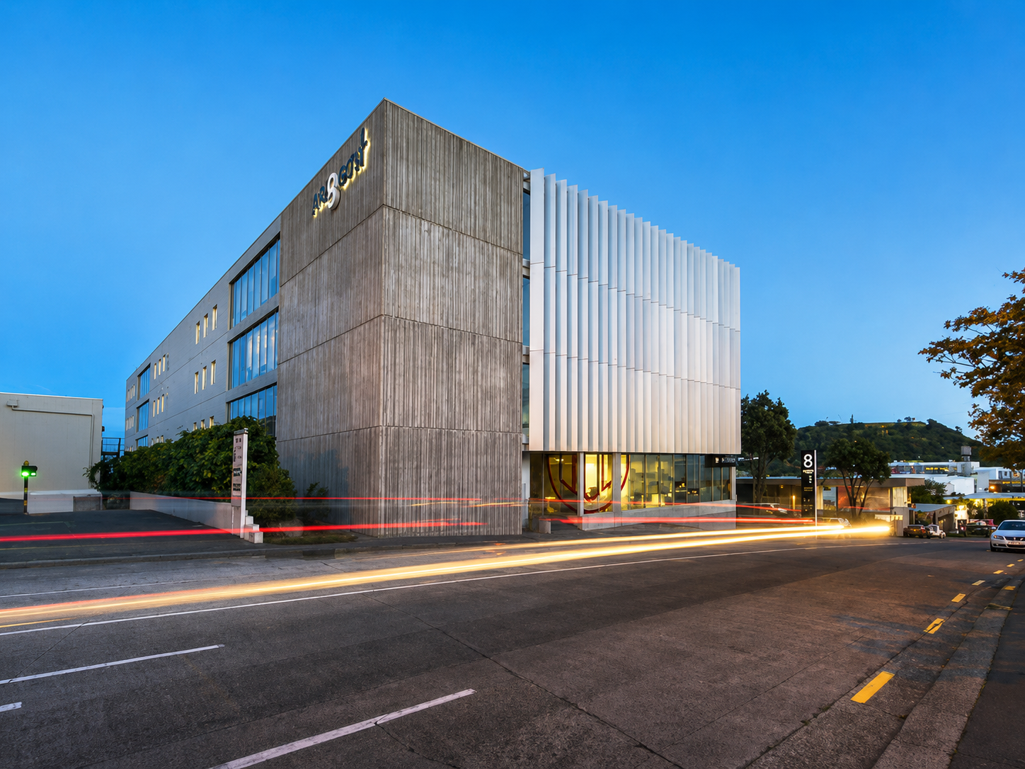 Modern multi-story office building with concrete and white panel facade at dusk with light trails on the street.