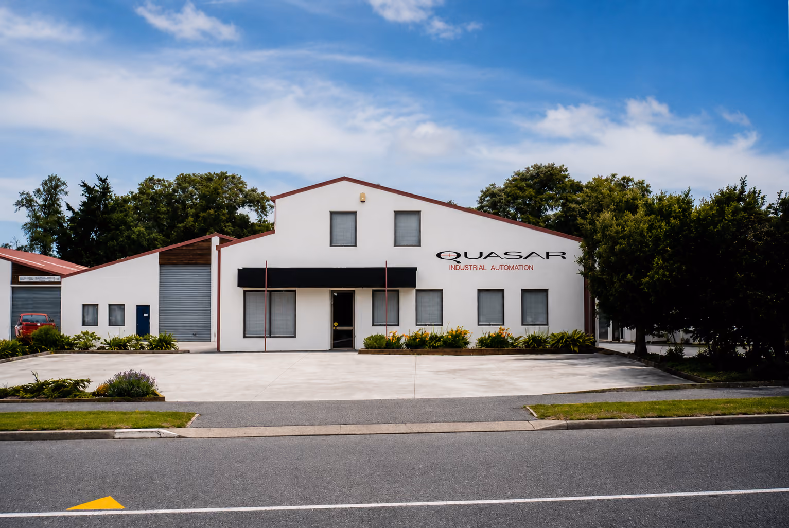 White industrial building with QUASAR INDUSTRIAL AUTOMATION sign and parking in front under blue sky.