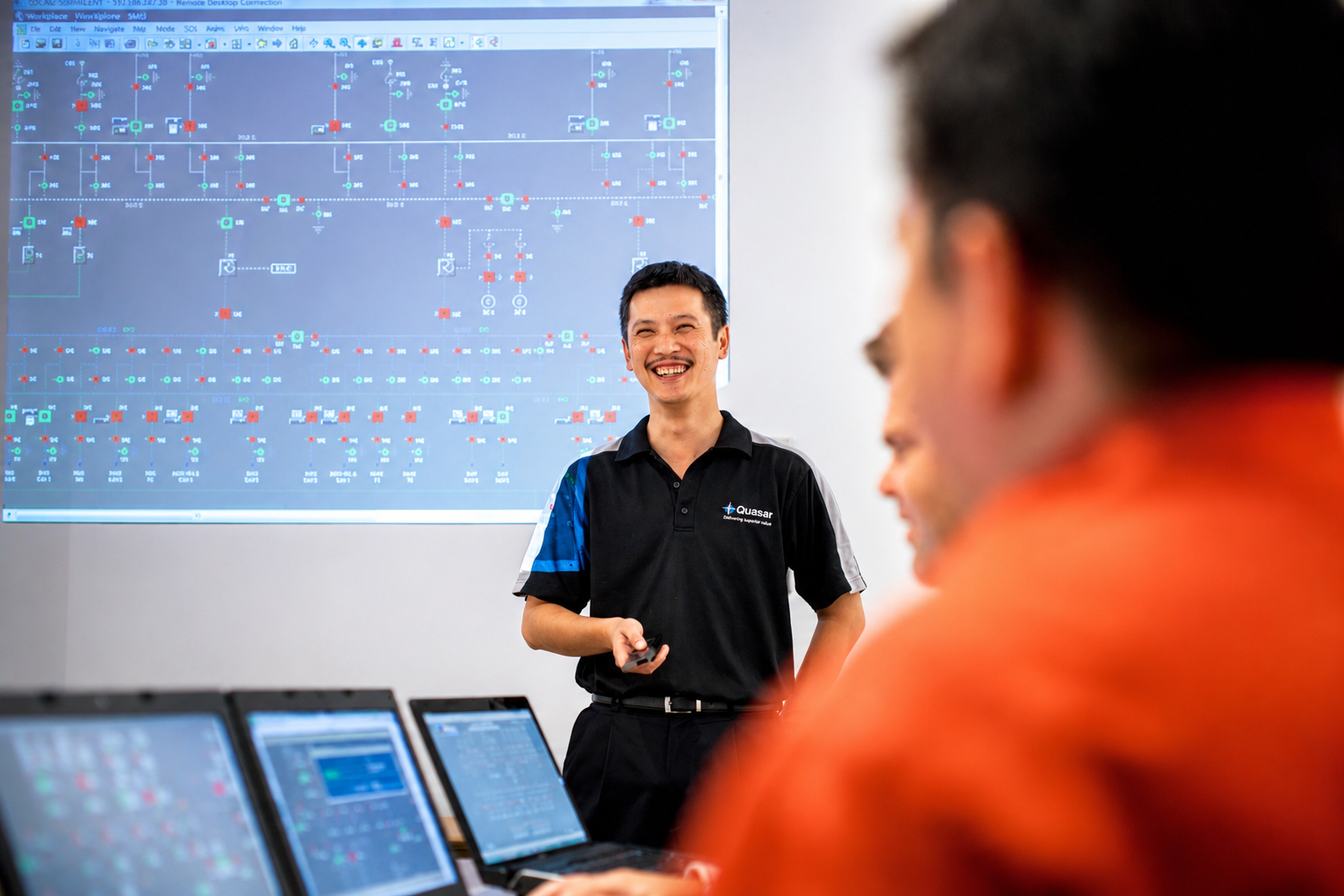Man in Quasar shirt smiling and presenting in front of a screen with technical data and laptops.