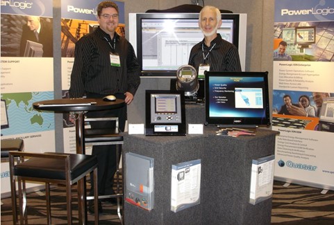 Two men standing at a technology booth with monitors and electronic devices at a conference.