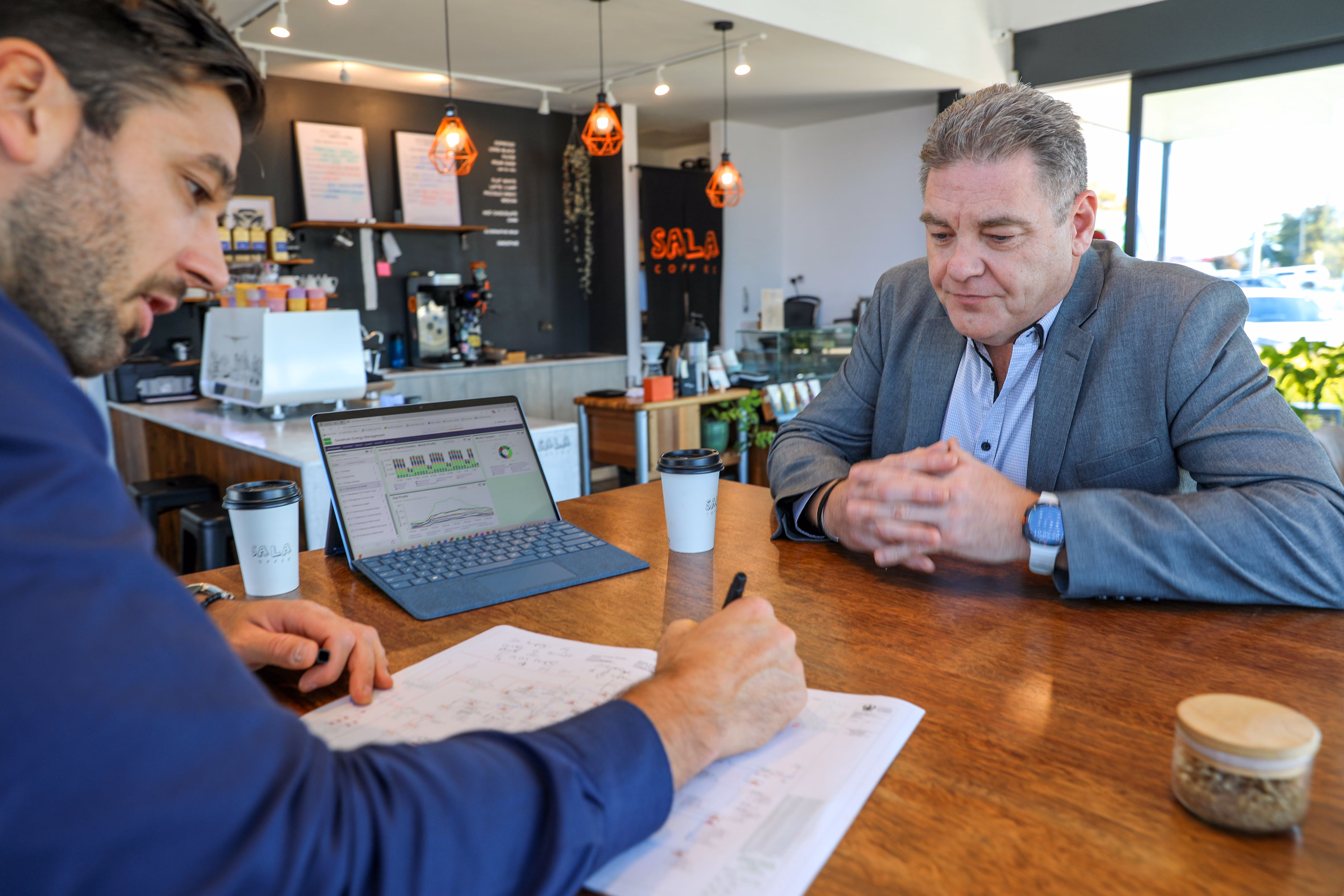 Two men at a wooden table reviewing documents with a laptop displaying energy data charts.