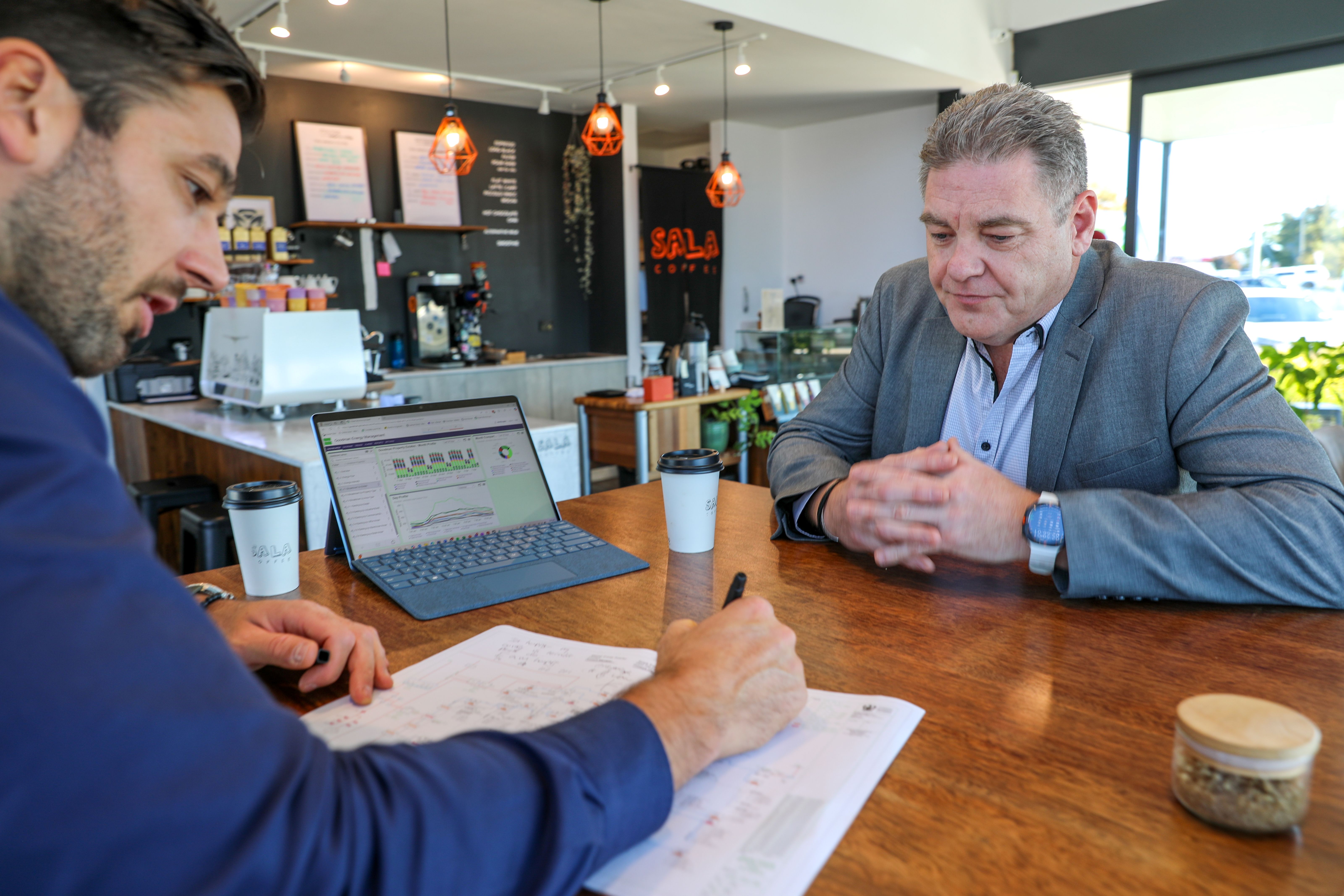 Two men at a wooden table reviewing documents with a laptop displaying energy data charts.