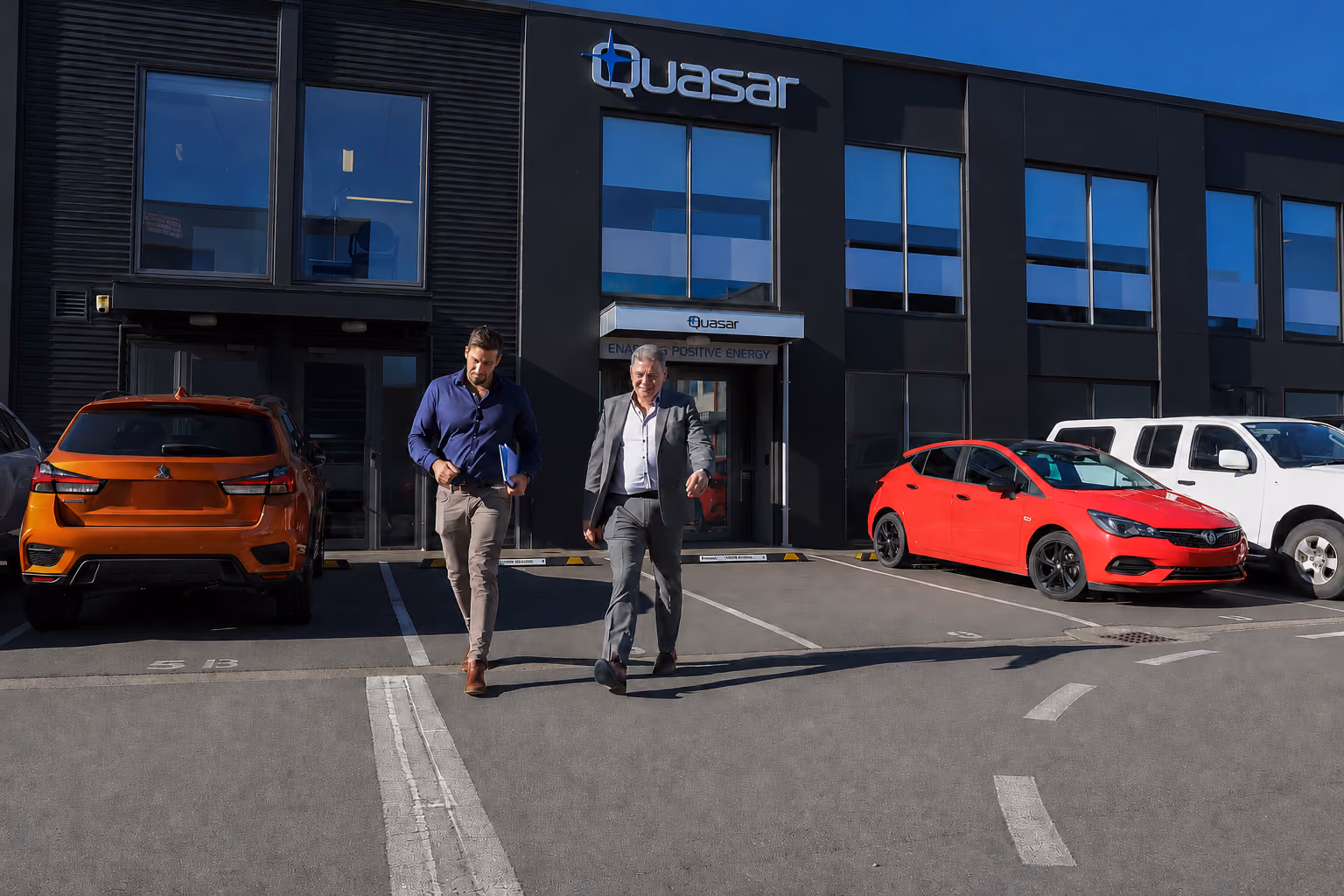 Two men walking in front of a black Quasar office building with cars parked outside.