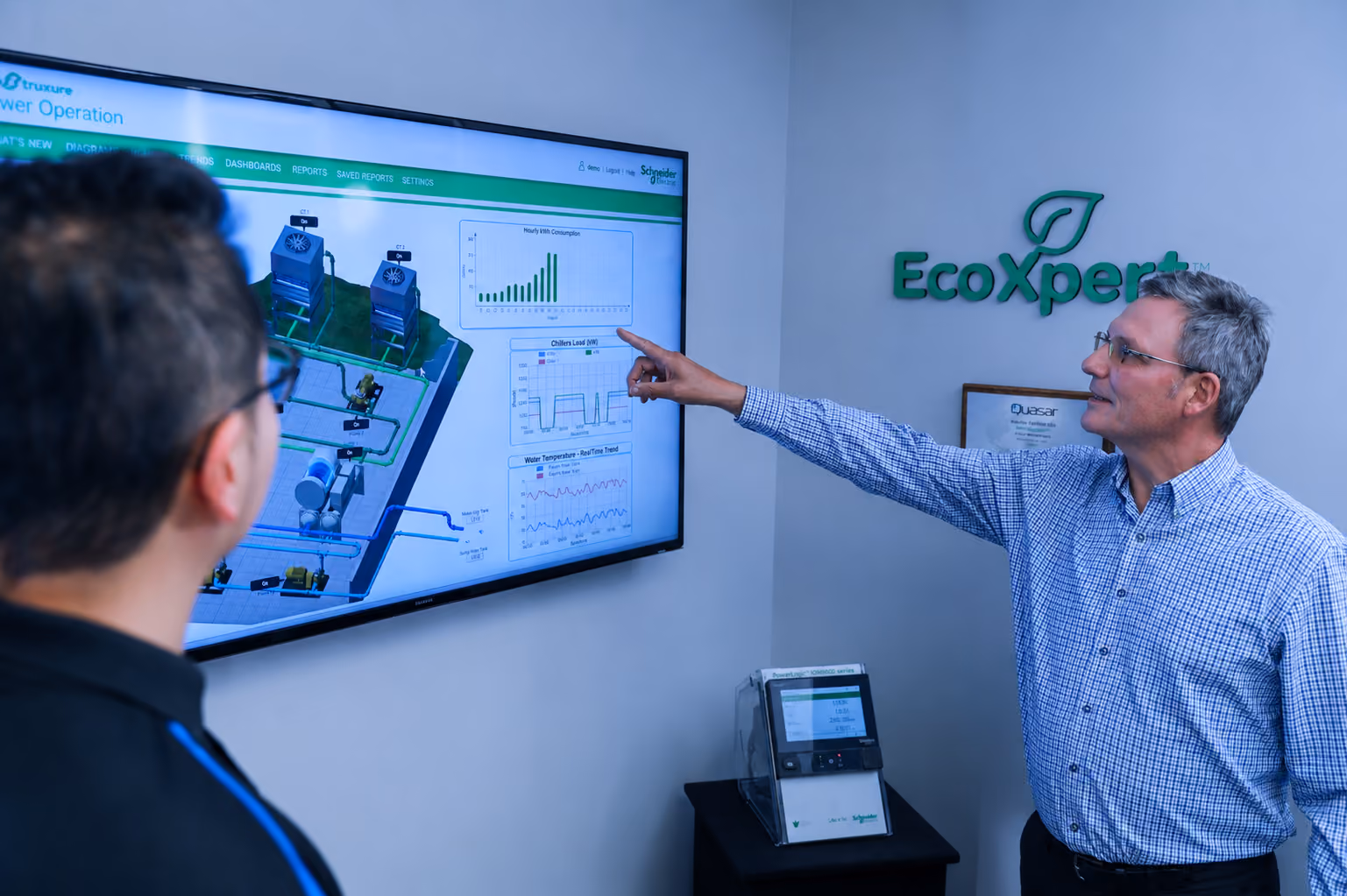 Older man points at data charts on wall screen while younger man watches in an office with EcoXpert sign.