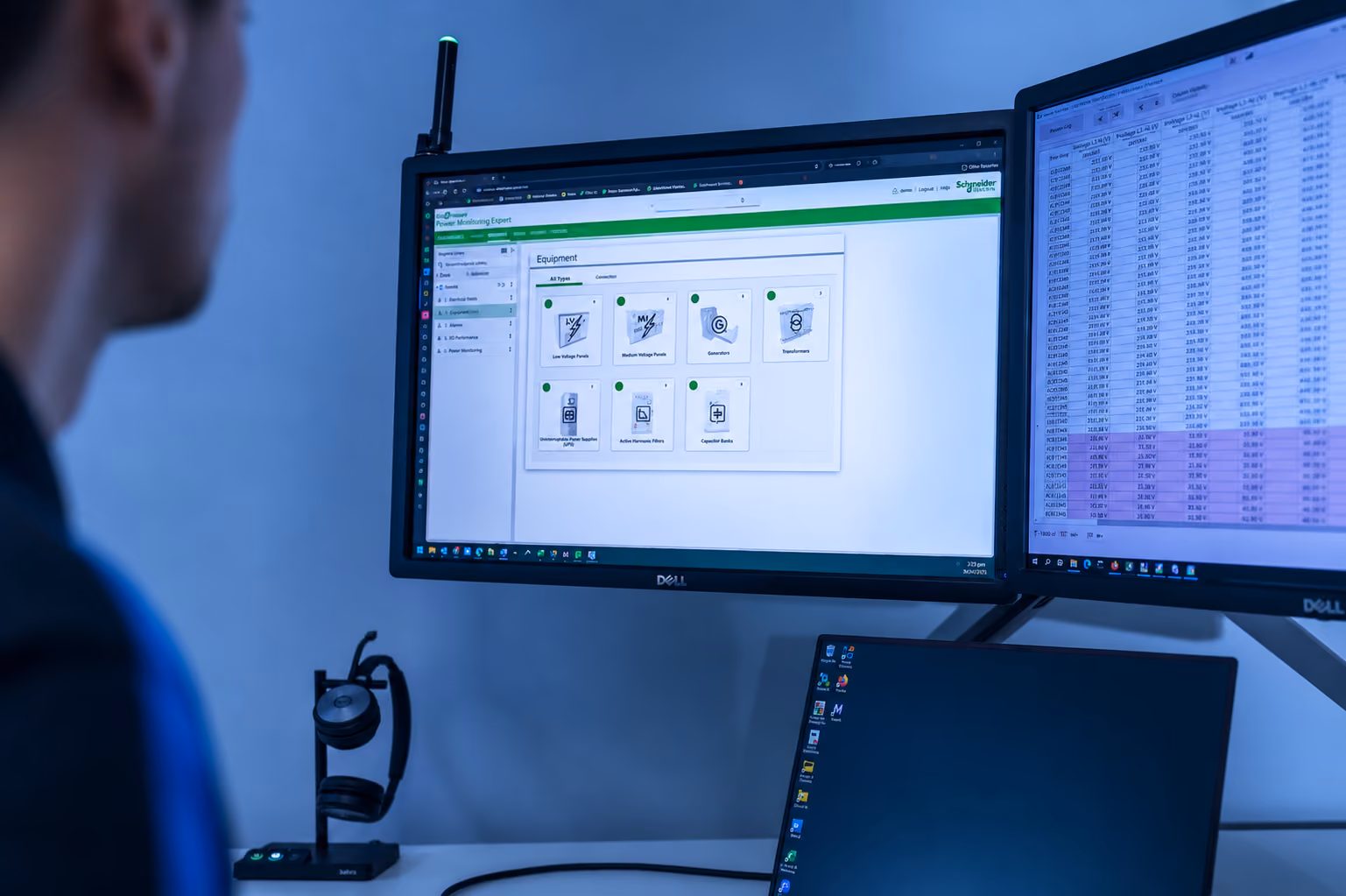 Person views dual monitors showing equipment data and voltage readings with a headset on the desk.