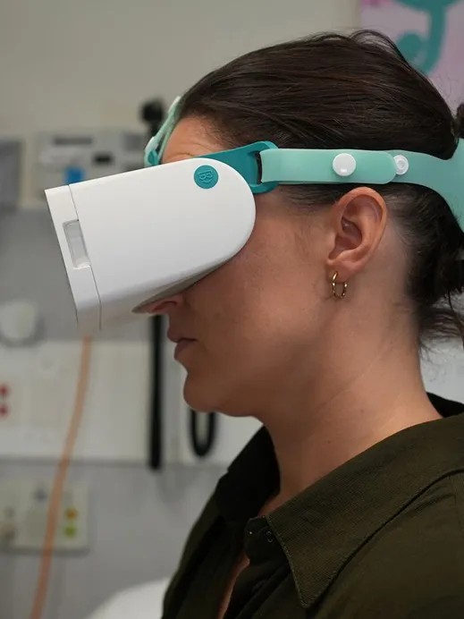Side view of a woman wearing a white and teal virtual reality headset in a clinical or office setting.