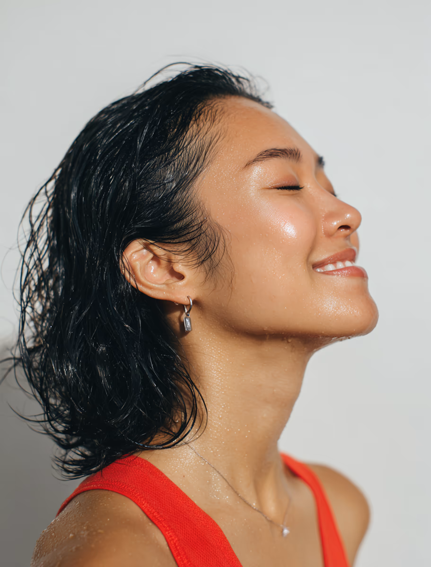 Side profile of a smiling woman with wet hair wearing a red top, with water droplets on her skin.