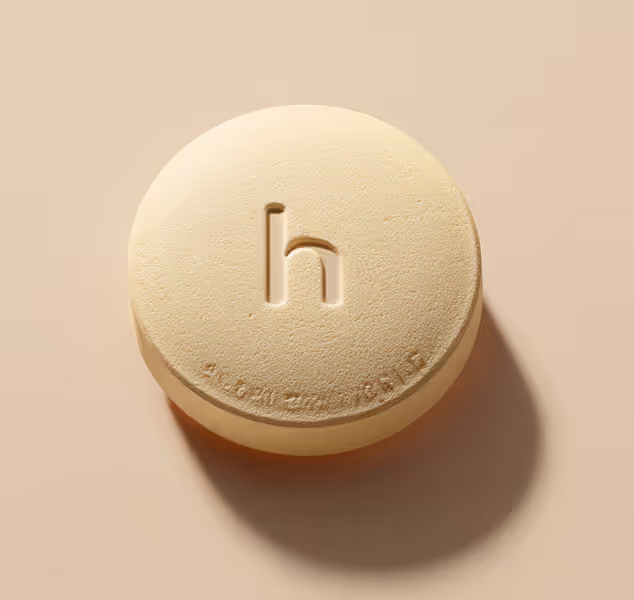 Round beige pill with a lowercase 'h' engraved on top and small text along the edge, casting a shadow on a beige surface.
