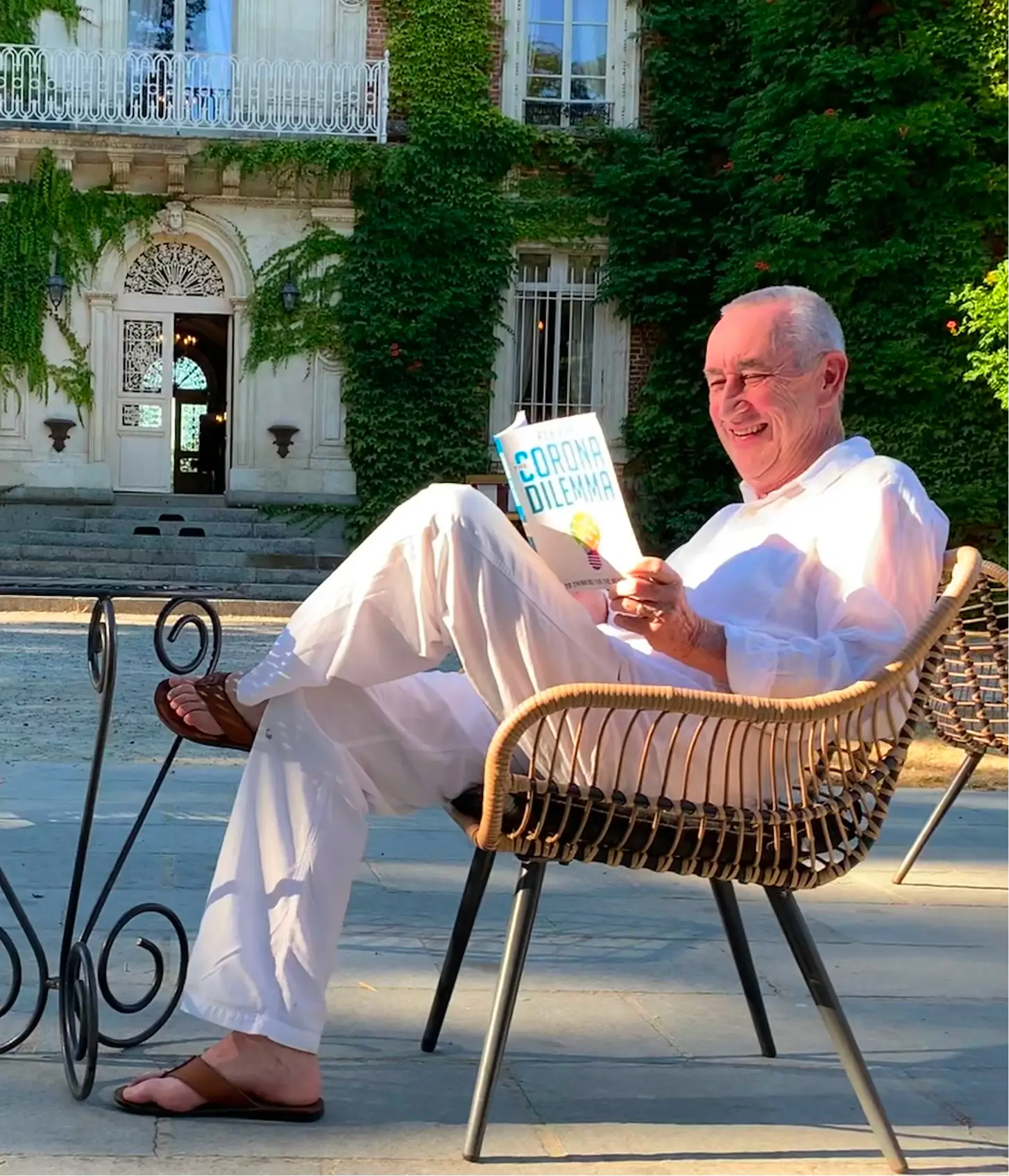 Smiling elderly man in white clothing sits outdoors on a wicker chair reading a book titled 'Dream Today What You Can Achieve Tomorrow'.