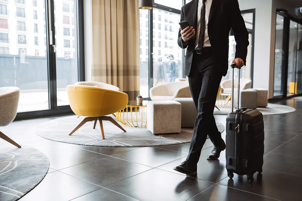 Man in a black suit walking through a modern hotel lobby pulling a black suitcase and holding a smartphone.