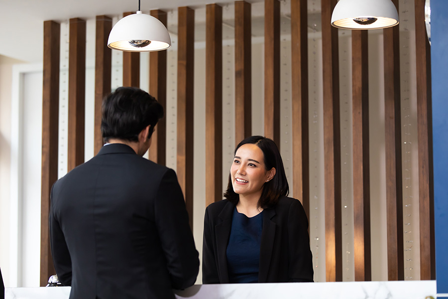 Receptionist smiling and talking to a male guest at a hotel front desk with modern wooden panel background.