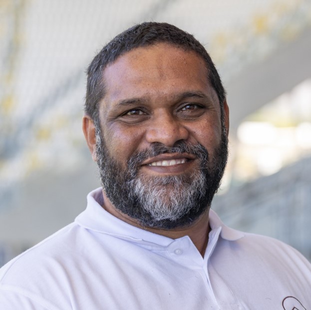 Man with short curly hair and a beard smiling, wearing a white collared shirt, with a blurred indoor background.