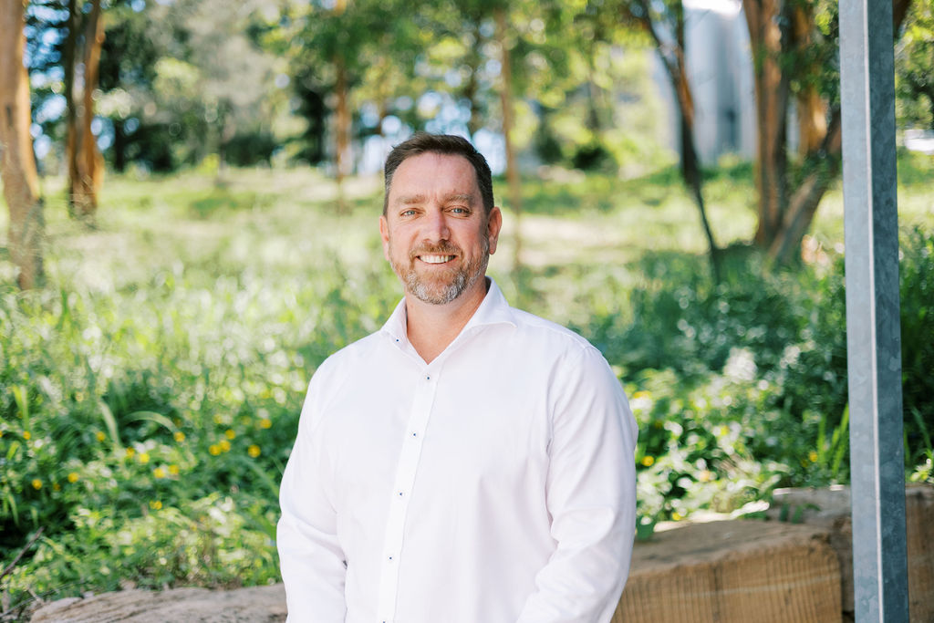 Smiling man with short dark hair and beard wearing a white button-up shirt standing outdoors with greenery in the background.
