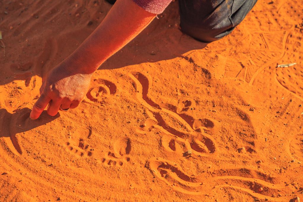 Hand drawing an abstract animal figure in reddish sand using a finger.