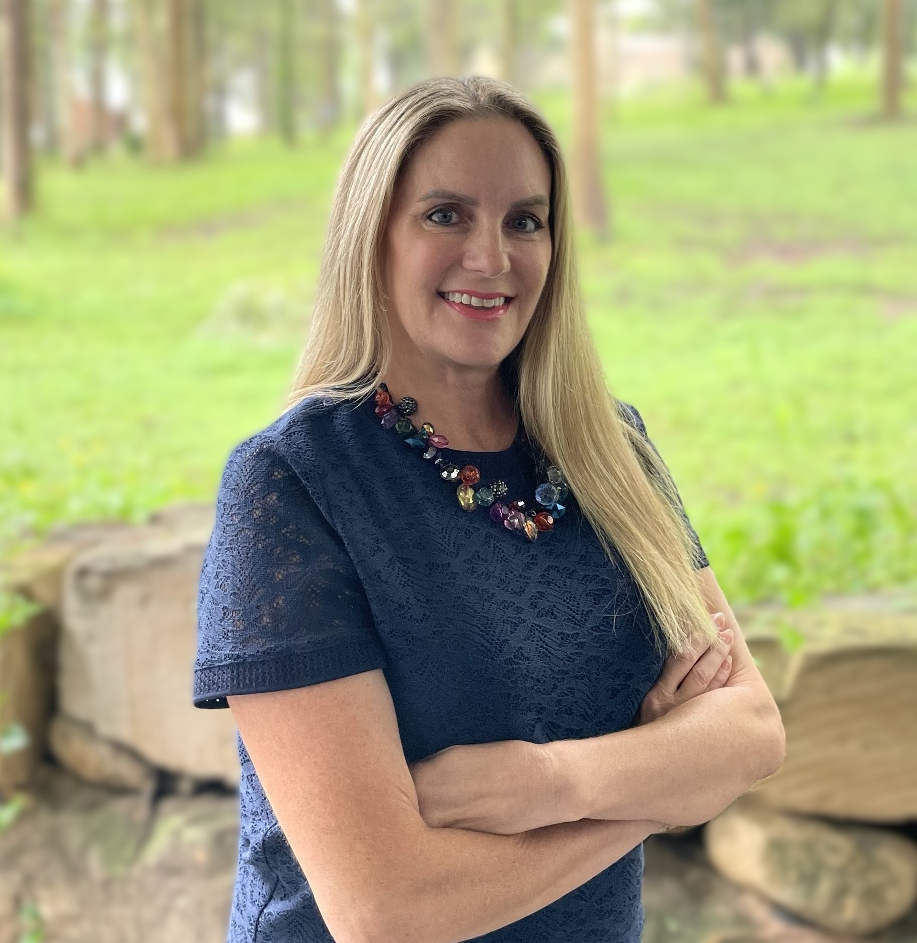 Smiling woman with long blonde hair in a navy lace top and colorful beaded necklace standing outdoors with arms crossed.
