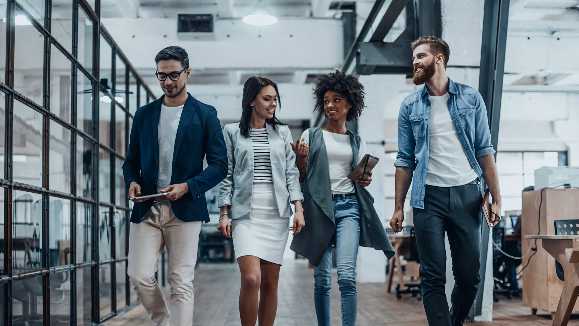 Four diverse young professionals walking and talking in a modern open office space.