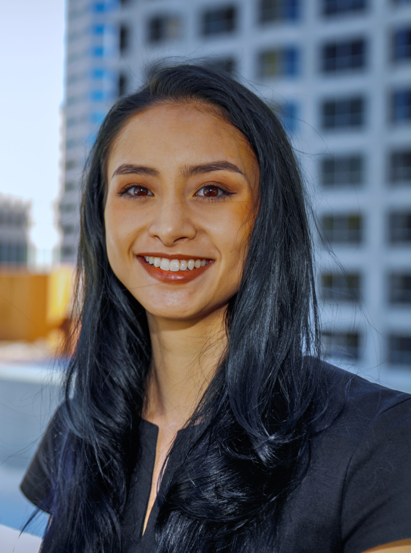 Smiling woman with long black hair standing outdoors in front of an urban building.