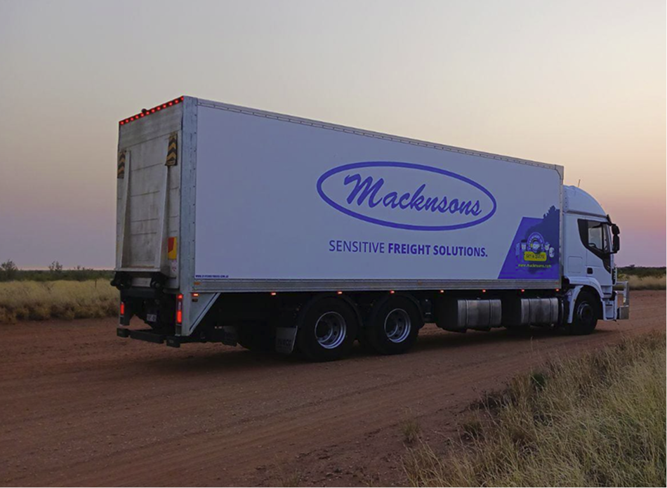 White Macknsons freight truck parked on a dirt road at sunset with grass and sparse trees in the background.