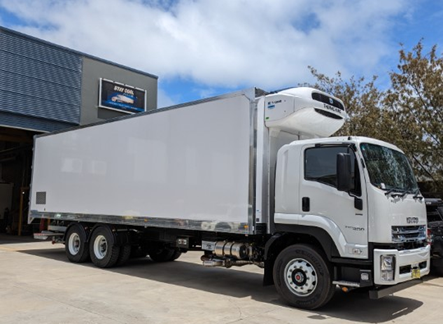 White refrigerated Isuzu truck parked on a concrete surface with a partly cloudy sky and trees in the background.