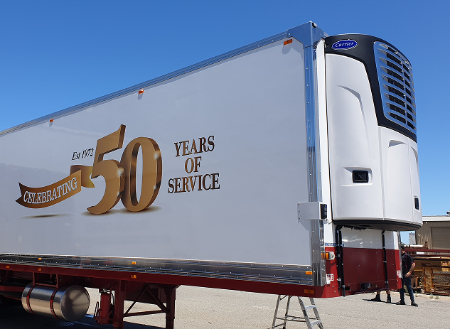Side view of a white refrigerated semi-trailer with 'Celebrating 50 years of service Est. 1972' printed on it, parked outdoors under a clear blue sky.