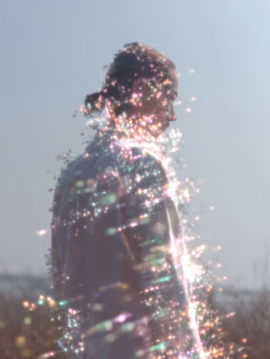 A soft-focus, ethereal portrait of a person in profile, their form shimmering with a dense cluster of glowing, multi-colored light particles and bokeh flares against a bright, hazy sky.