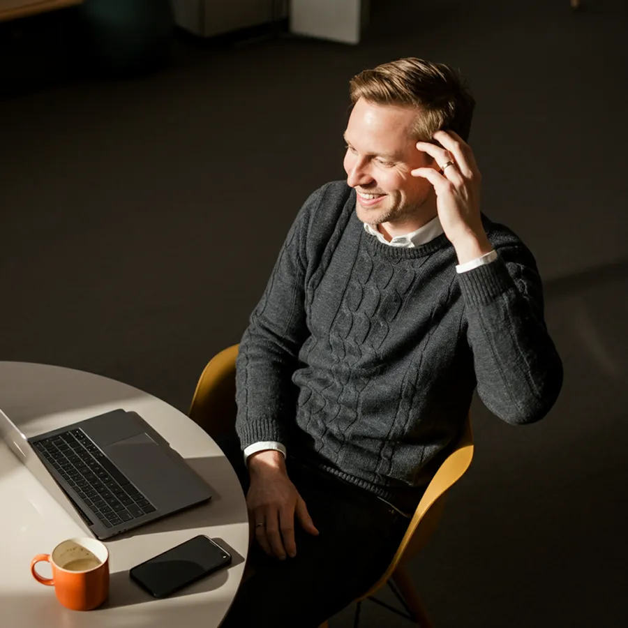 Ein lächelnder Mann in einem grauen Pullover sitzt an einem Tisch mit einem Laptop, Smartphone und einer orangefarbenen Kaffeetasse.