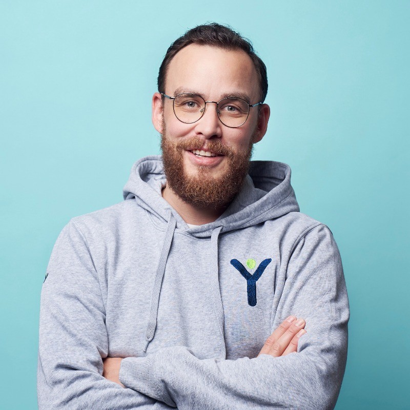 Lächelnder Mann mit Bart und Brille trägt grauen Hoodie mit aufgesticktem Y-Logo vor hellblauem Hintergrund.