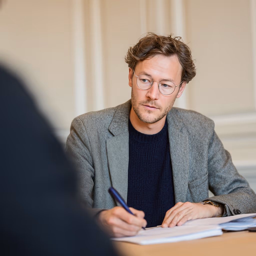 Mann mit Brille und grauem Blazer, der an einem Tisch sitzt und Notizen auf einem Dokument macht.