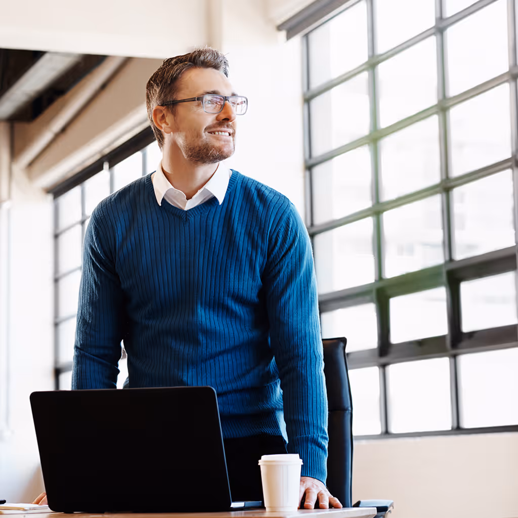Ein Mann mit Brille und blauem Pullover steht lächelnd an einem Schreibtisch mit Laptop und Kaffeebecher vor großen Fenstern.