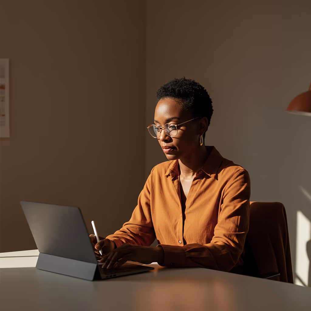 Frau mit Brille und braunem Hemd arbeitet konzentriert an einem Laptop mit Stylus in einem ruhigen, sonnendurchfluteten Raum.
