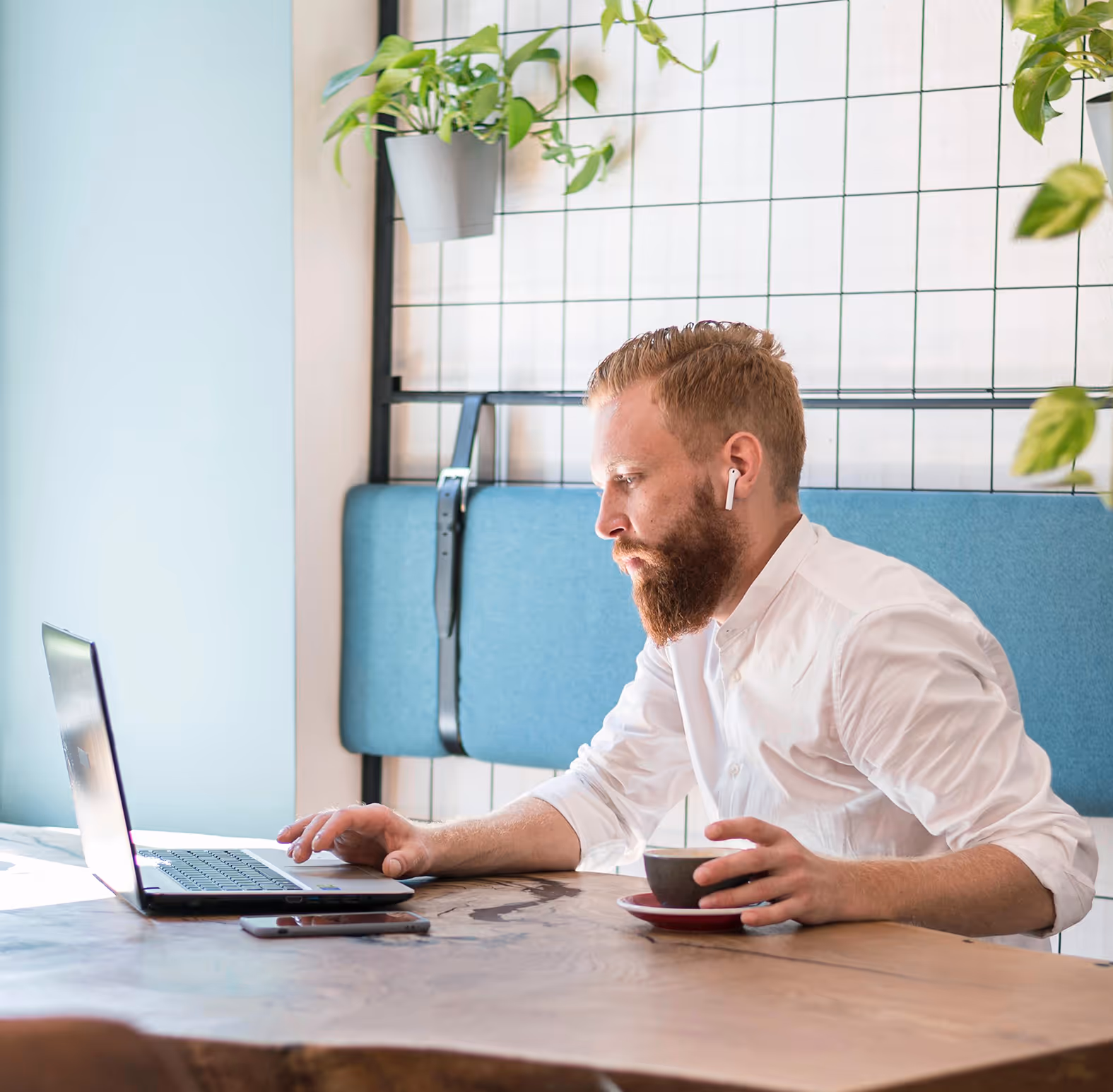 Seitlichaufnahme eines bärtigen Mannes mit weißen Kopfhörern, der an einem Laptop in einem modernen Raum mit Pflanzen arbeitet und eine Tasse hält.