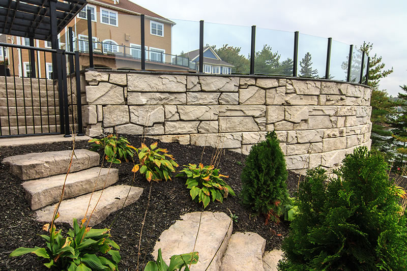 Outdoor stone staircase leading up beside a curved stone retaining wall with glass panels on top and landscaping with shrubs and plants.