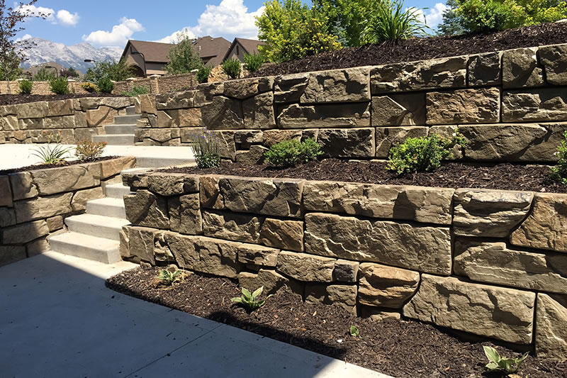 Sunlit tiered retaining wall made of large stone blocks with small plants and mulch, concrete steps leading up, and houses and mountains in the background.
