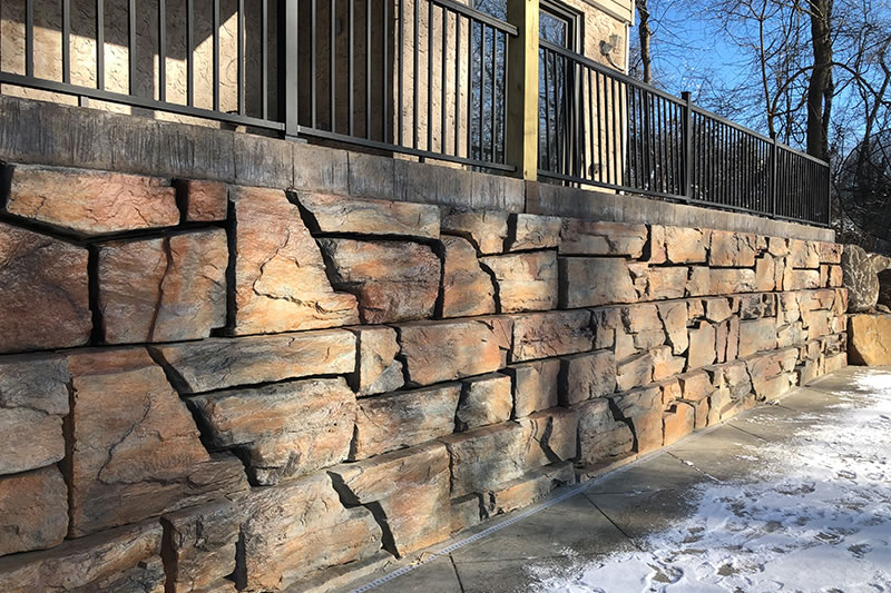 Stone retaining wall with large, irregularly shaped brownish stones next to a concrete pathway partially covered in snow, with a metal railing above.