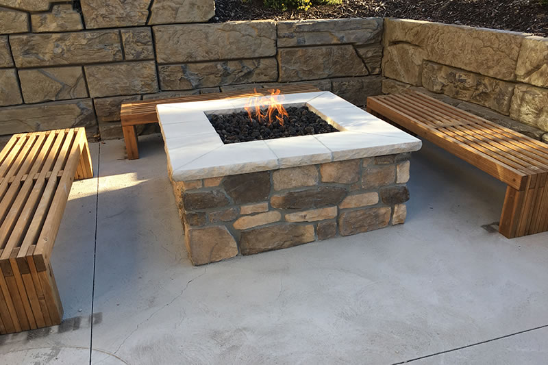 Stone fire pit with small flames surrounded by three wooden benches on a concrete patio with stone retaining walls.