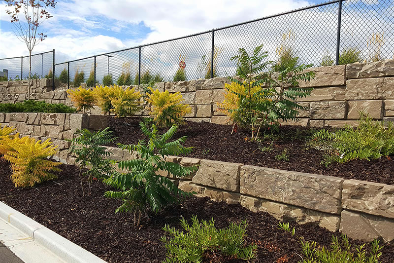 Tiered stone retaining wall with mulched garden beds featuring green and yellow shrubs under a partly cloudy sky.