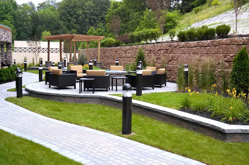 Outdoor patio area with cushioned black chairs and tables, surrounded by stone retaining walls, greenery, and a pergola in the background.