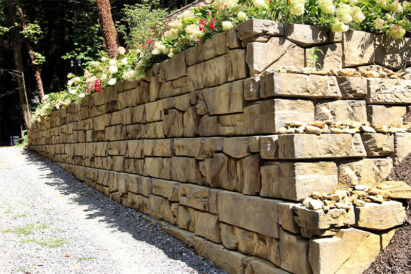 Stone retaining wall with layered large rectangular blocks topped with flowering plants along a gravel path.