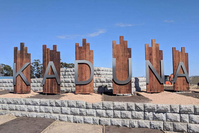 Large metal letters spelling 'KADUNA' mounted on tall rustic wooden panels, surrounded by stone retaining walls under a clear blue sky.