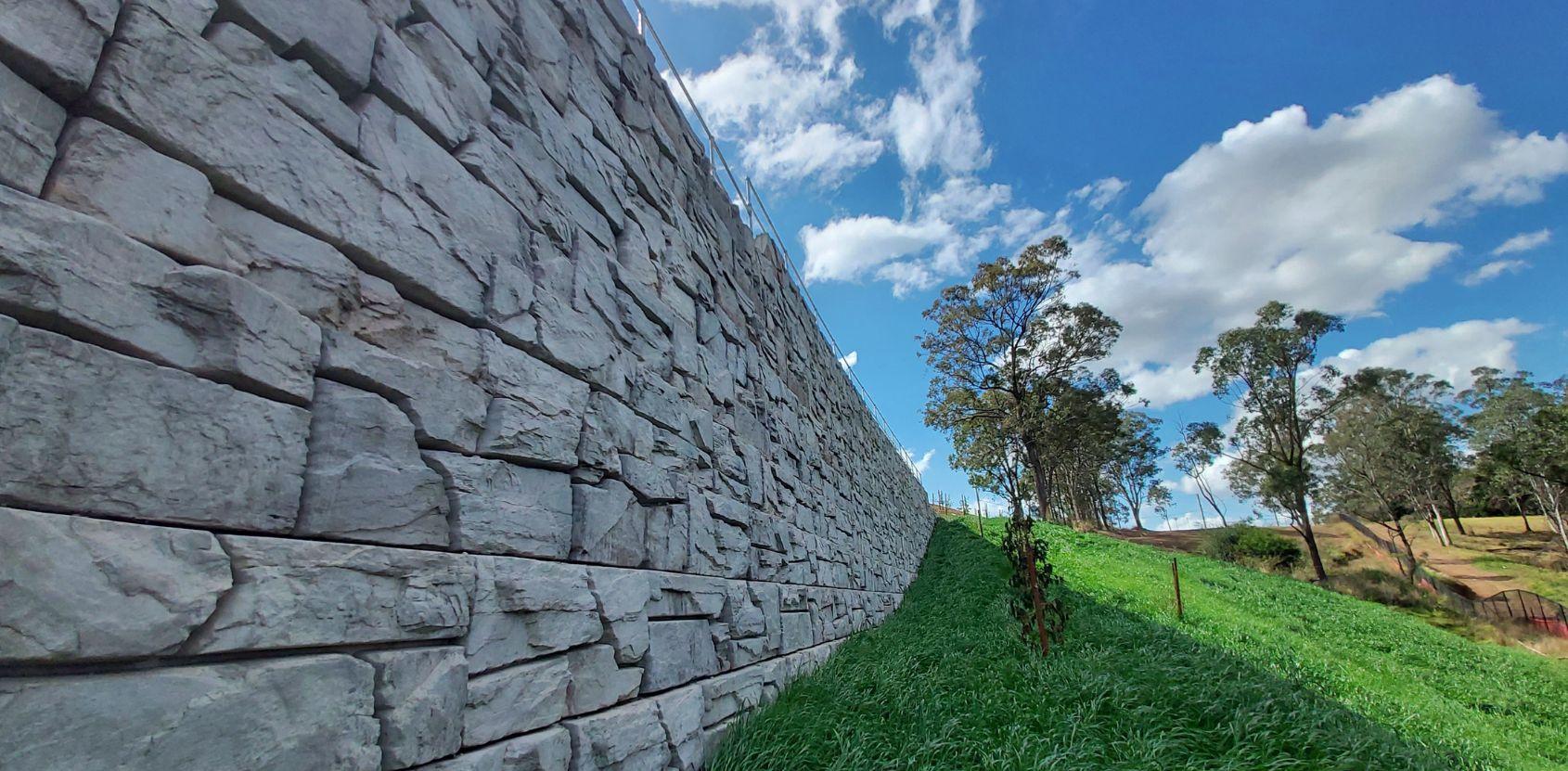 Tall stone retaining wall next to a grassy slope with scattered trees under a partly cloudy blue sky.