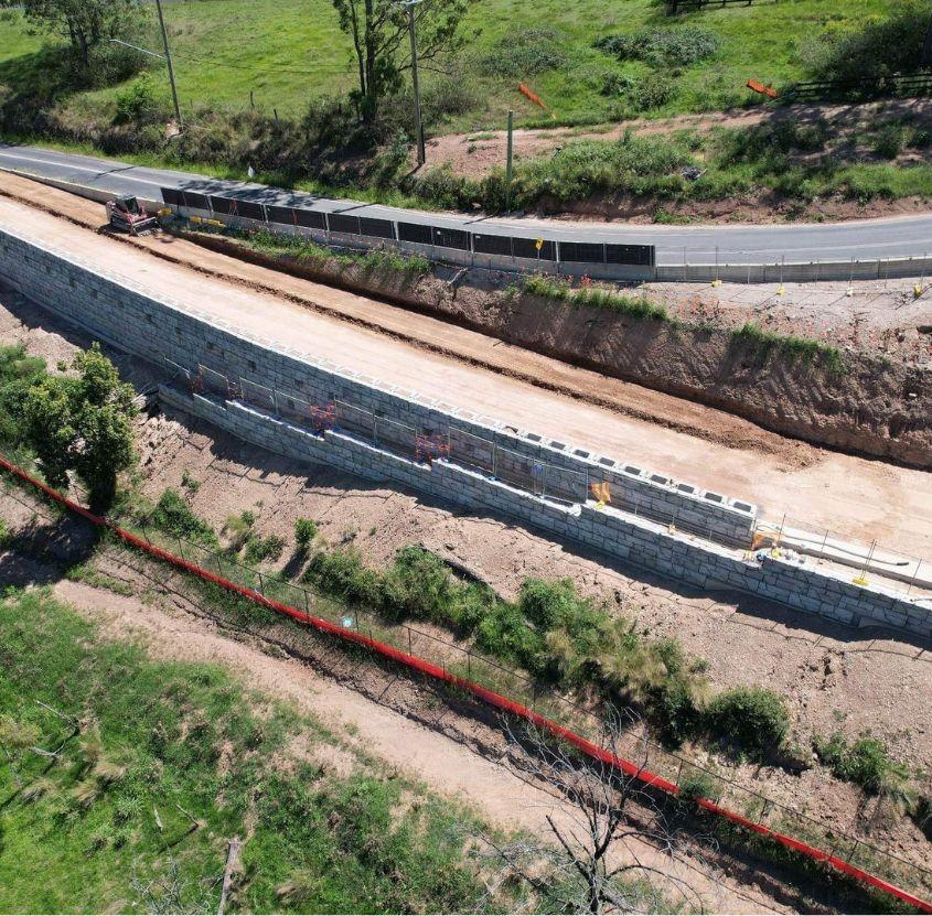 Long retaining wall under construction next to a road with green grassy hill in the background.