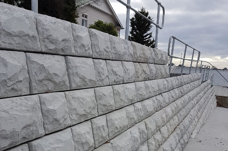 Angled view of a textured concrete retaining wall with metal handrails, adjacent to a house and a tree under a cloudy sky.