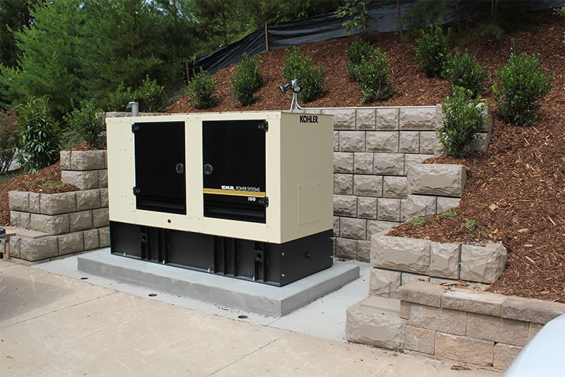 Kohler power system generator installed on a concrete pad next to a retaining wall and landscaped hillside.