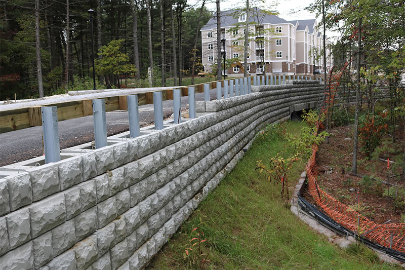 Curved retaining wall made of concrete blocks along a road near trees and a residential building.