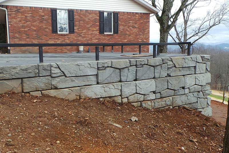 Stone retaining wall with gray rectangular blocks in front of a brick house with black window shutters.