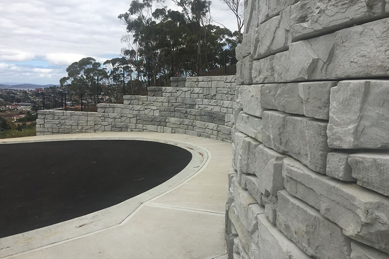 Curved stone retaining wall along a concrete sidewalk with blacktop road and trees in the background under a cloudy sky.