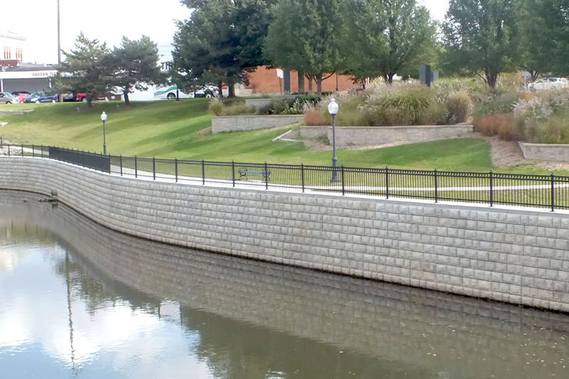 A stone retaining wall with black metal railing along a calm river, with a grassy park and trees in the background.