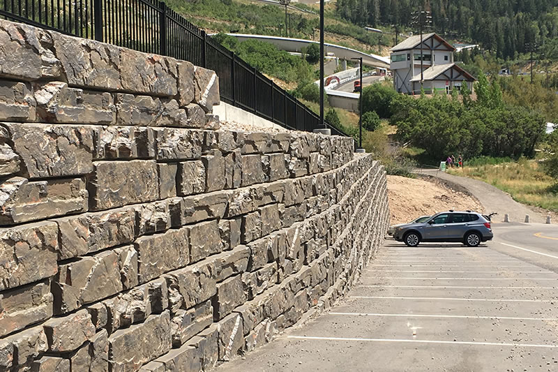 Stone retaining wall next to a parking lot with a single blue SUV parked and a house with hills and greenery in the background.