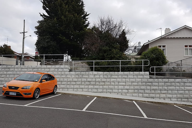 Orange car parked in an empty lot next to a tiered stone retaining wall with metal railings, trees, and a house in the background under a cloudy sky.